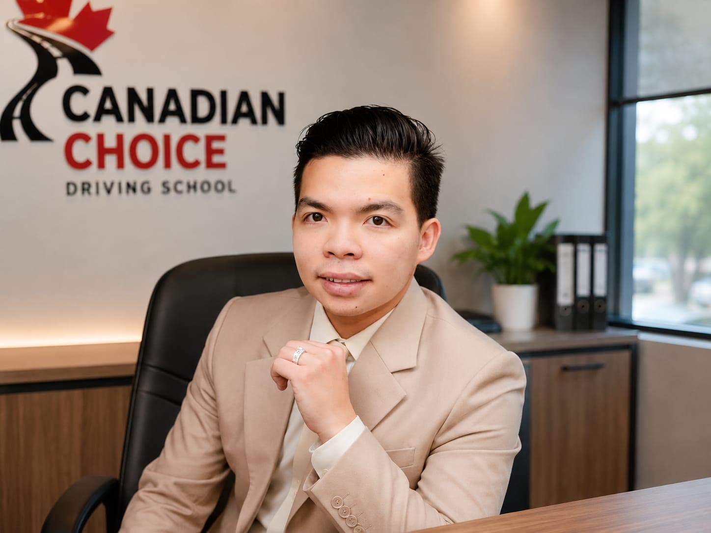 Duong, Huu Thanh, driving instructor at Canadian Choice Driving School, seated in a beige suit at a desk in the school's office in front of the Canadian Choice Driving School wall logo.